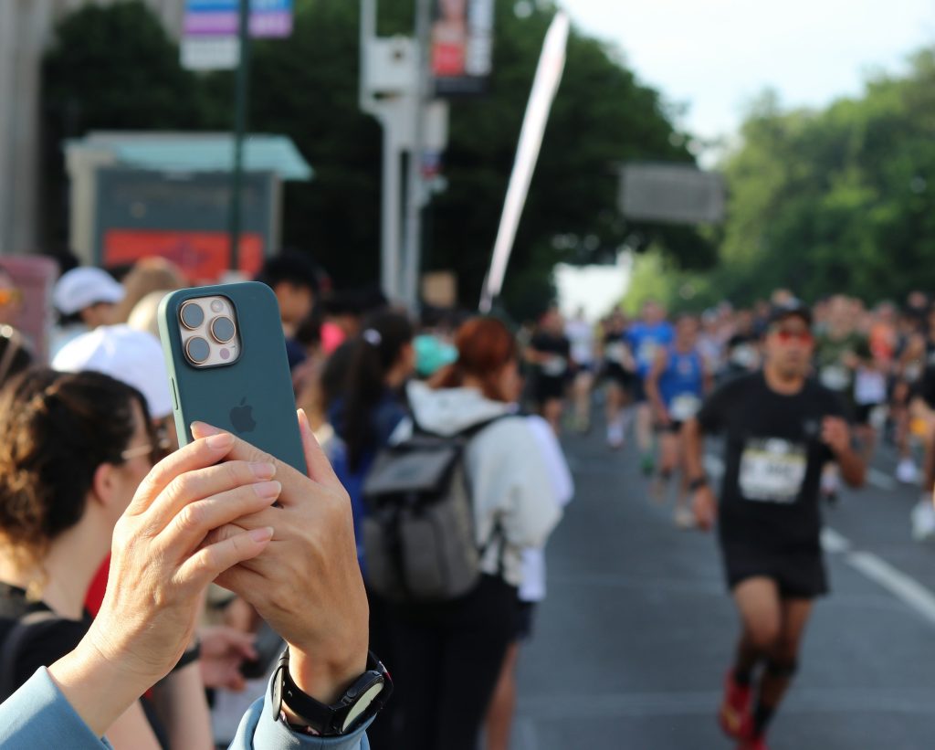 Marathon spectator holding a phone, with crowd behind and runners passing on the right