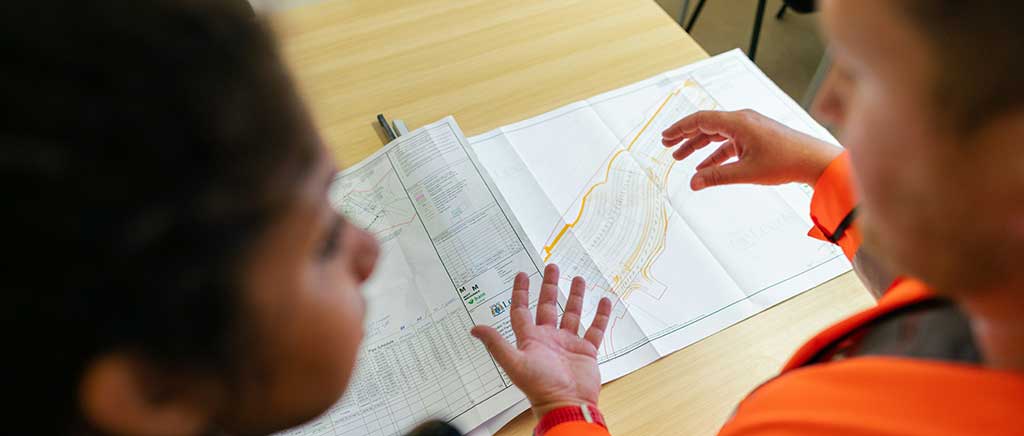 Two people, one in high-vis, in discussion with an architect's building plan and building data charts.