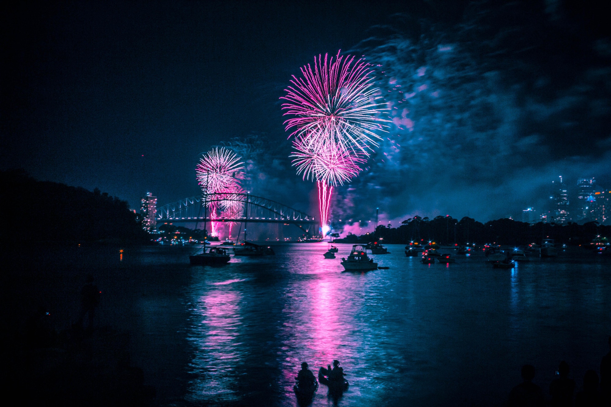 Pink and white fireworks above a dark blue city scene, rivers on a boat are shown in the foreground