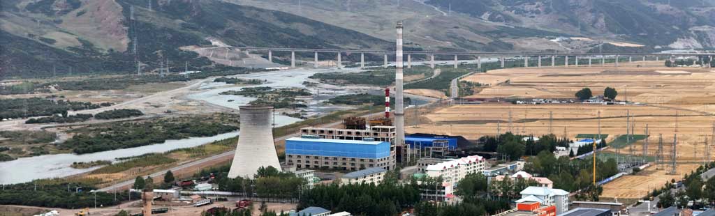 Cooling tower, factory and residential buildings, next to a river and fields, with roads and pylons behind.