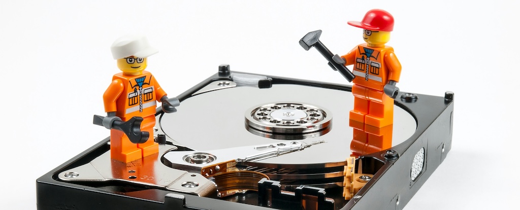 A disk drive, removed from a computer, on a white background. On top are two lego figures dressed as engineers holding tools. 